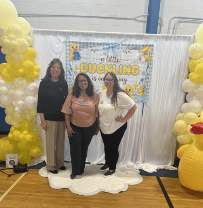 A row of three people in front of a duckling-themed display with yellow balloons.